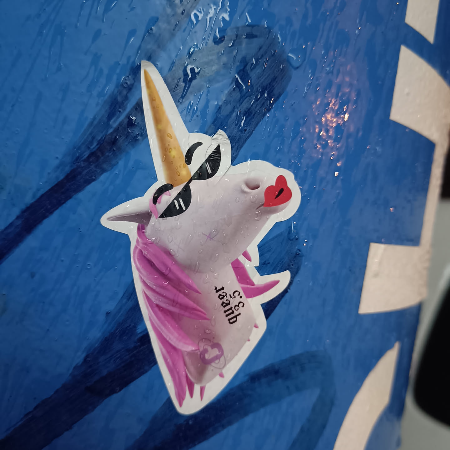 A sticker of a unicorn wearing sunglasses and lipstick. The unicorn's horn is gold, and its mane is pink. There is some text on the sticker that says 'C&C 18amb'.
