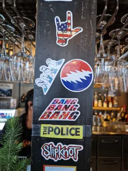 A close-up shot of several stickers on a dark surface.  The central sticker features the band The Police's name in yellow with the words "Bang Bang Bang" stacked above it in a stylized font. Other stickers include a Grateful Dead steal your face logo, Slipknot logo, and a hand making the 'I love you' sign with an American flag design.