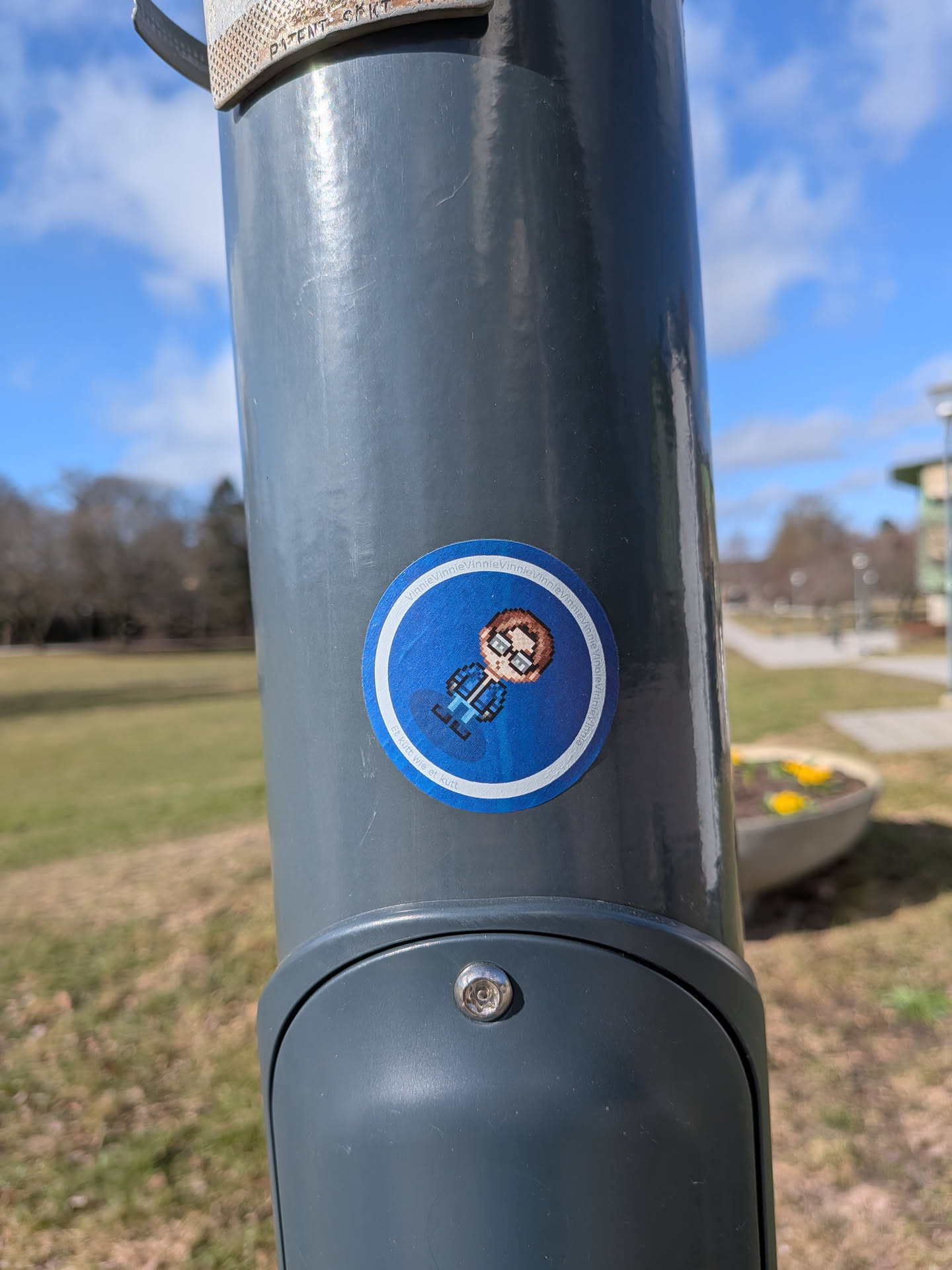 A circular sticker featuring a pixel art character against a vibrant blue background. The character is a male figure with brown hair, light skin, wearing glasses, a blue jacket, and dark pants/shoes. The art style is distinctly 8-bit or pixelated. A thin white border separates the blue background from the outer edge. Around the top inner edge of the blue circle, the name 'Vinnie' is repeated multiple times. Below the character, there is a phrase in what appears to be German.