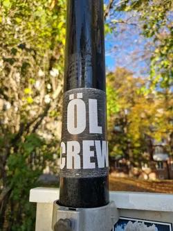 A black and white sticker with the text "ÖL CREW" on it is attached to a black pole. The sticker shows some wear and tear.