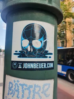A sticker featuring a skull design with a spiderweb pattern, melting into a puddle of liquid. The sticker has a website address and a smiley face logo. The colors are black, white, and light blue.
