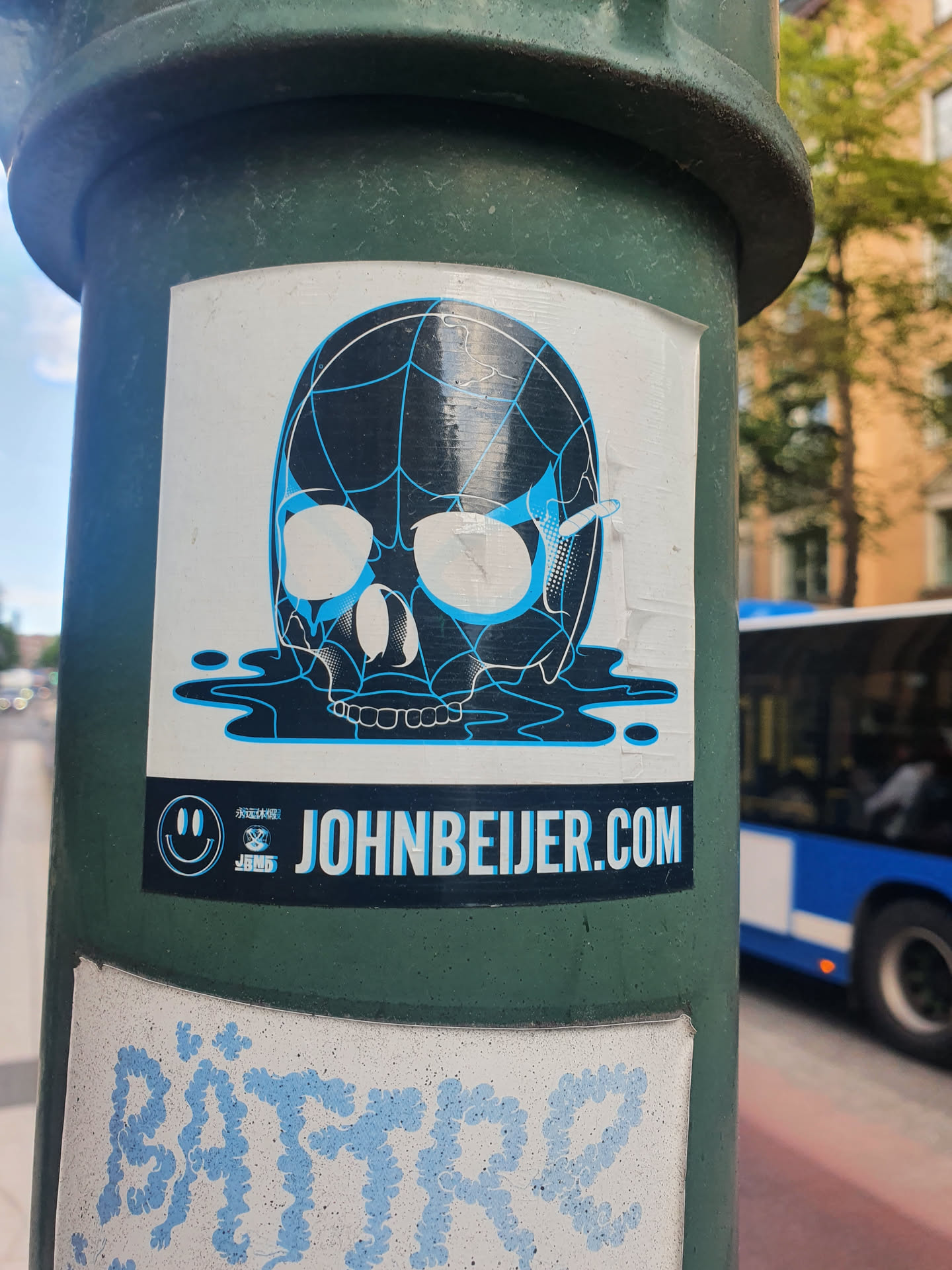 A sticker featuring a skull design with a spiderweb pattern, melting into a puddle of liquid. The sticker has a website address and a smiley face logo. The colors are black, white, and light blue.