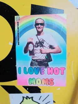 A sticker featuring a young, bald man with sunglasses holding a bottle of what seems to be a drink. He is depicted against a rainbow background. The text "I LOVE HOT MOMS" is written in colorful, bubble-like letters at the bottom.  An Instagram handle is visible on the side.
