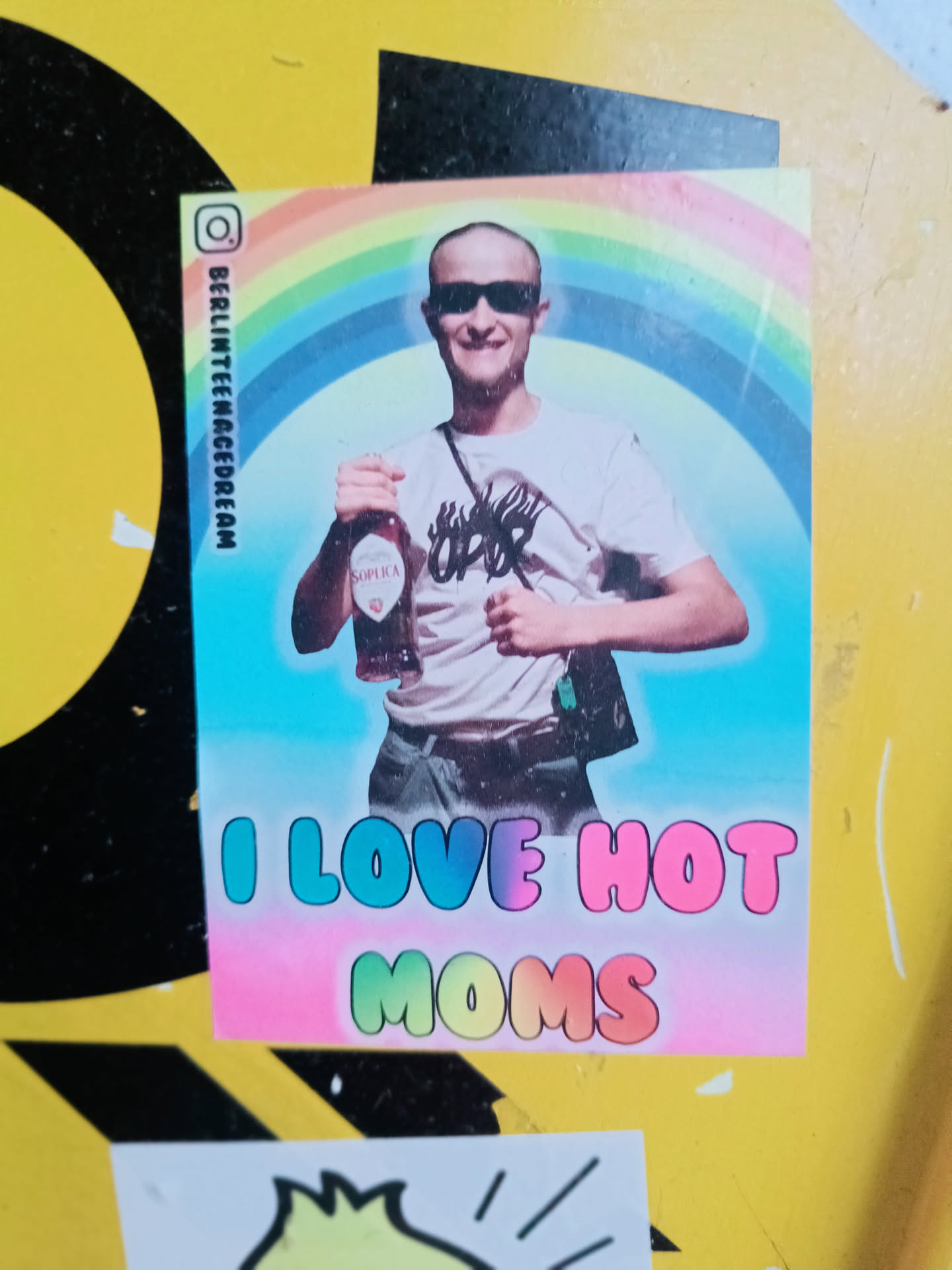 A sticker featuring a young, bald man with sunglasses holding a bottle of what seems to be a drink. He is depicted against a rainbow background. The text "I LOVE HOT MOMS" is written in colorful, bubble-like letters at the bottom.  An Instagram handle is visible on the side.