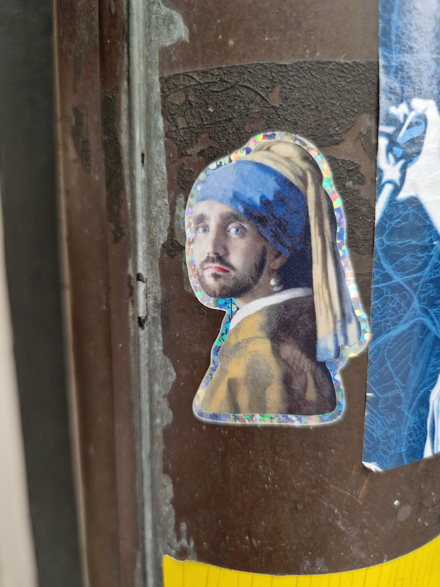 A holographic sticker featuring a modern interpretation of Johannes Vermeer's iconic "Girl with a Pearl Earring." The sticker is affixed to a brown, weathered surface, possibly a street pole or wall.