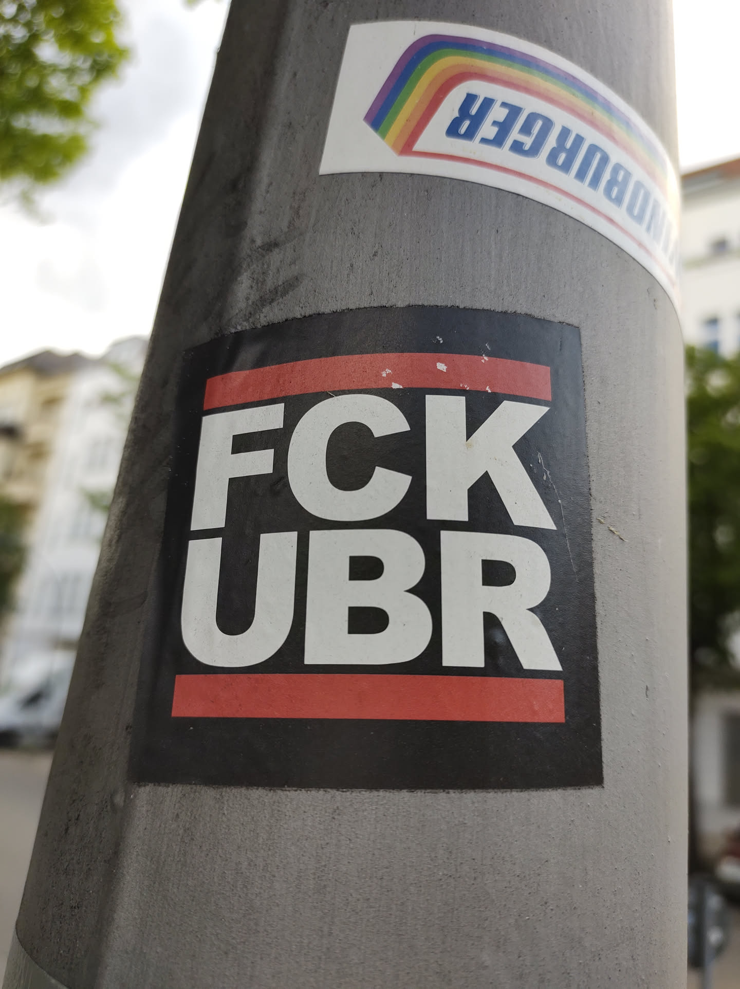 A black sticker with white and red text. The text says "FCk UBR" in bold uppercase letters. The sticker is affixed to a gray pole.