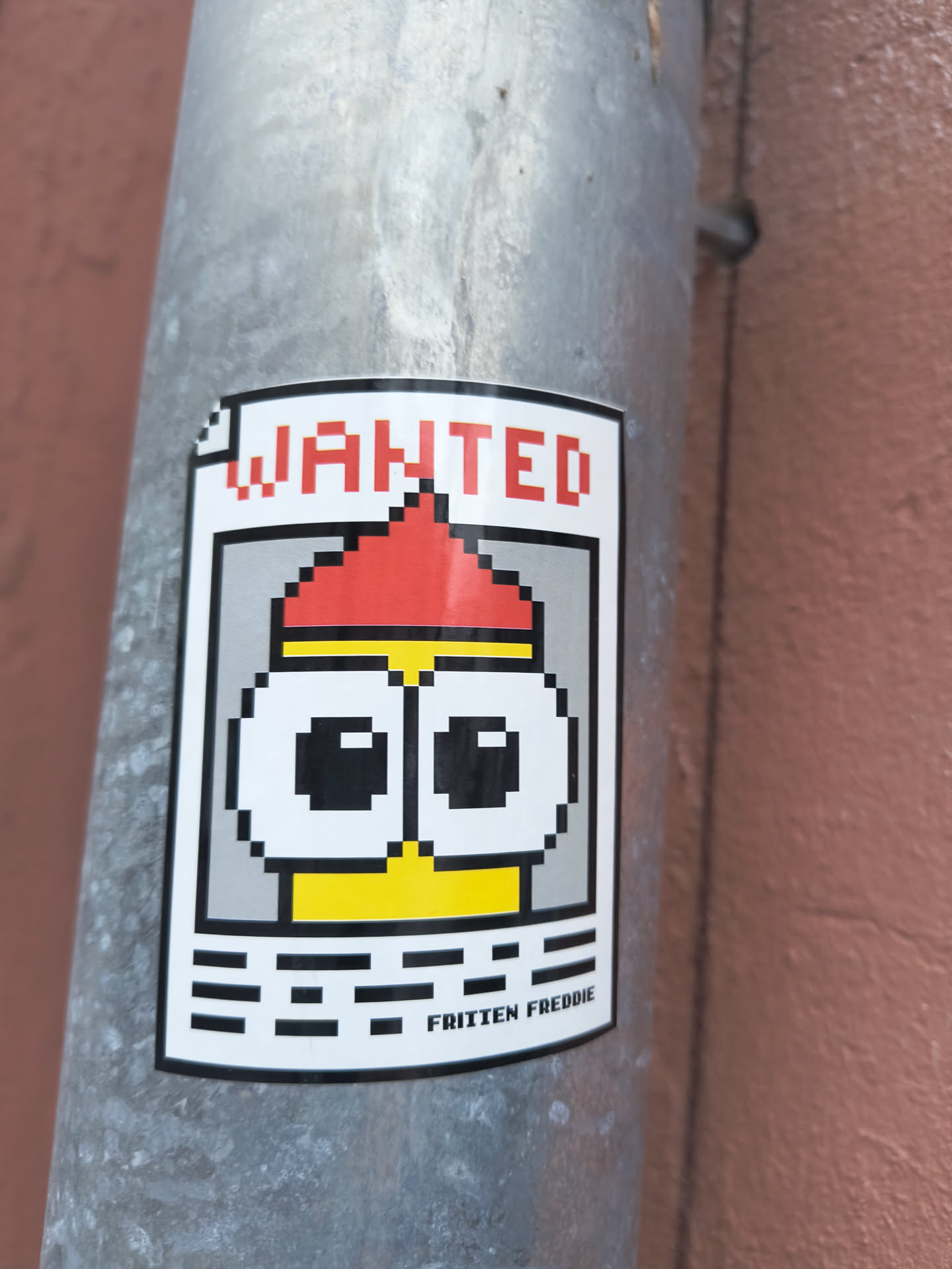 A rectangular sticker designed as a 'WANTED' poster, featuring a pixel art character. The sticker has a white background with a thick black border. At the top, the word 'WANTED' is written in bold, red, pixelated capital letters. Below this, a central character is depicted in a pixelated style, resembling a Minion-like figure. It has a yellow body/face, large white eyes with black pupils, and a red pointed hat or hair, separated by a thin yellow band. Below the character, there are several abstract horizontal black pixelated lines, suggesting a space for information. At the very bottom, centered, the name 'FRITTEN FREDDIE' is written in black, pixelated letters. The sticker is adhered to a textured, grey metallic pole, showing slight wear and a subtle curve.