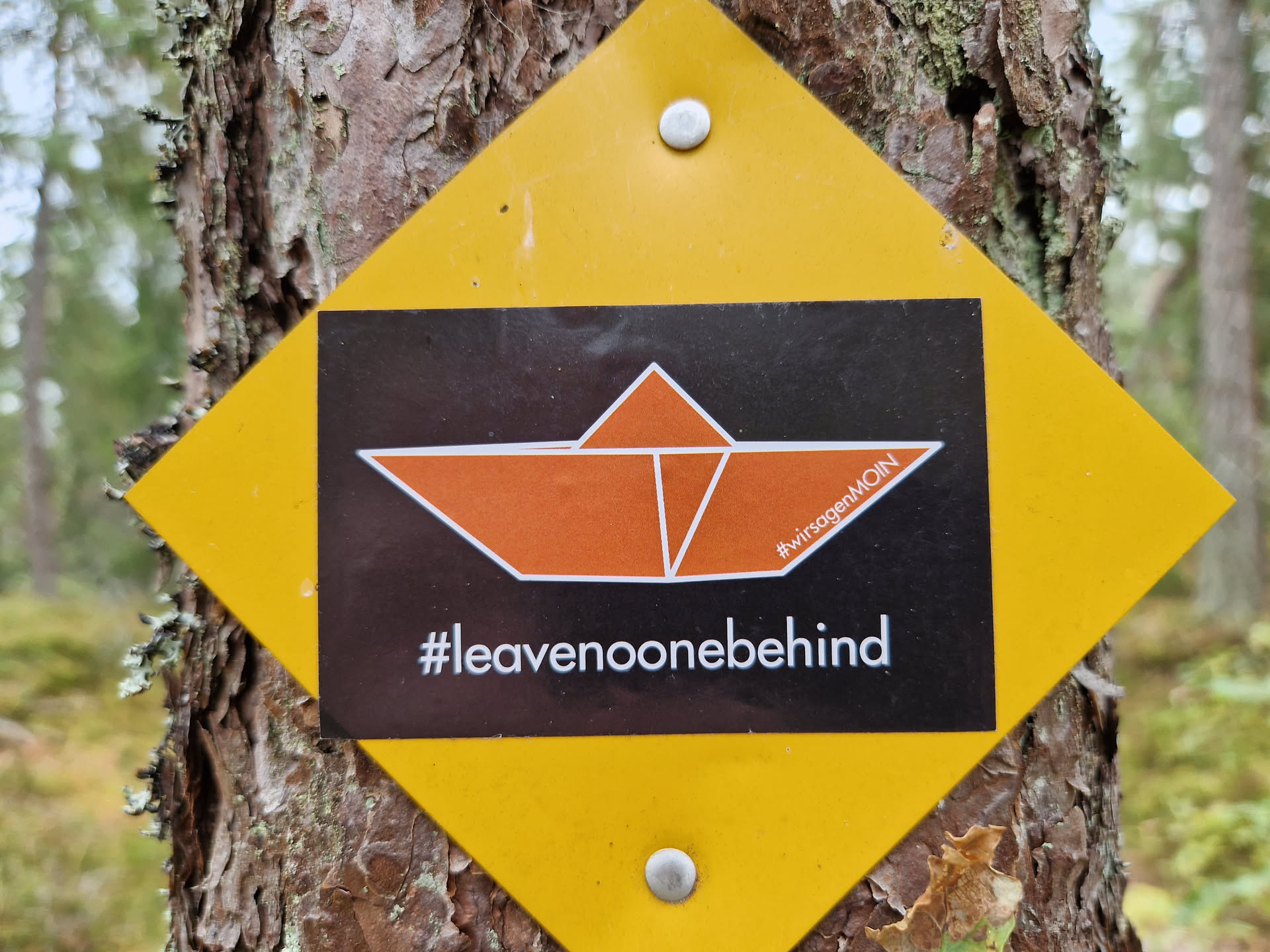 A yellow diamond-shaped sticker affixed to a tree. The sticker features a black background with an orange origami boat and the text '#leavenoonebehind' and '#wirborgenMOIN'.