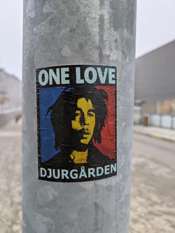 A sticker featuring Bob Marley's portrait with "One Love" and "Djurgården" text. The sticker is affixed to a metal pole.