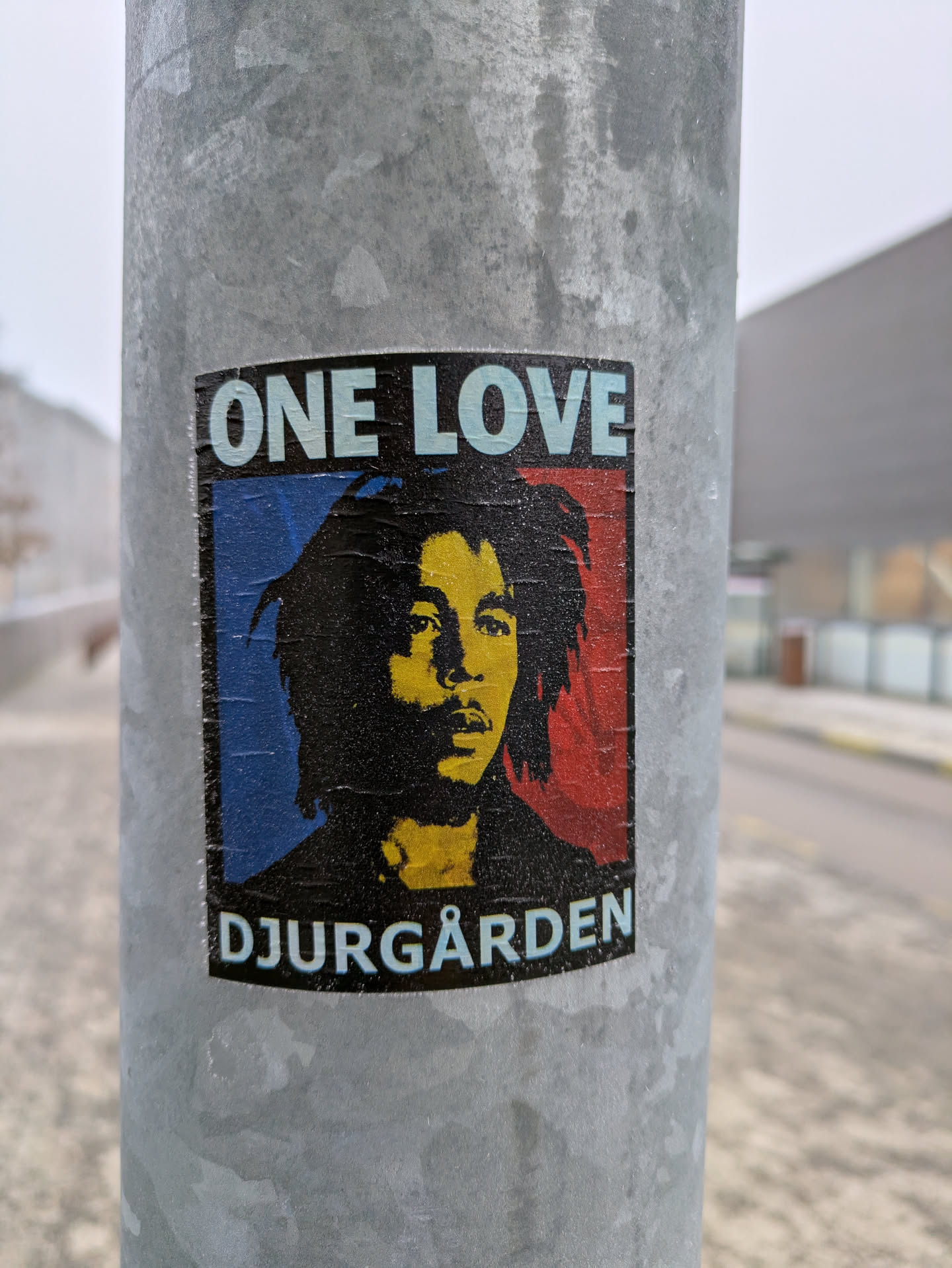 A sticker featuring Bob Marley's portrait with "One Love" and "Djurgården" text. The sticker is affixed to a metal pole.