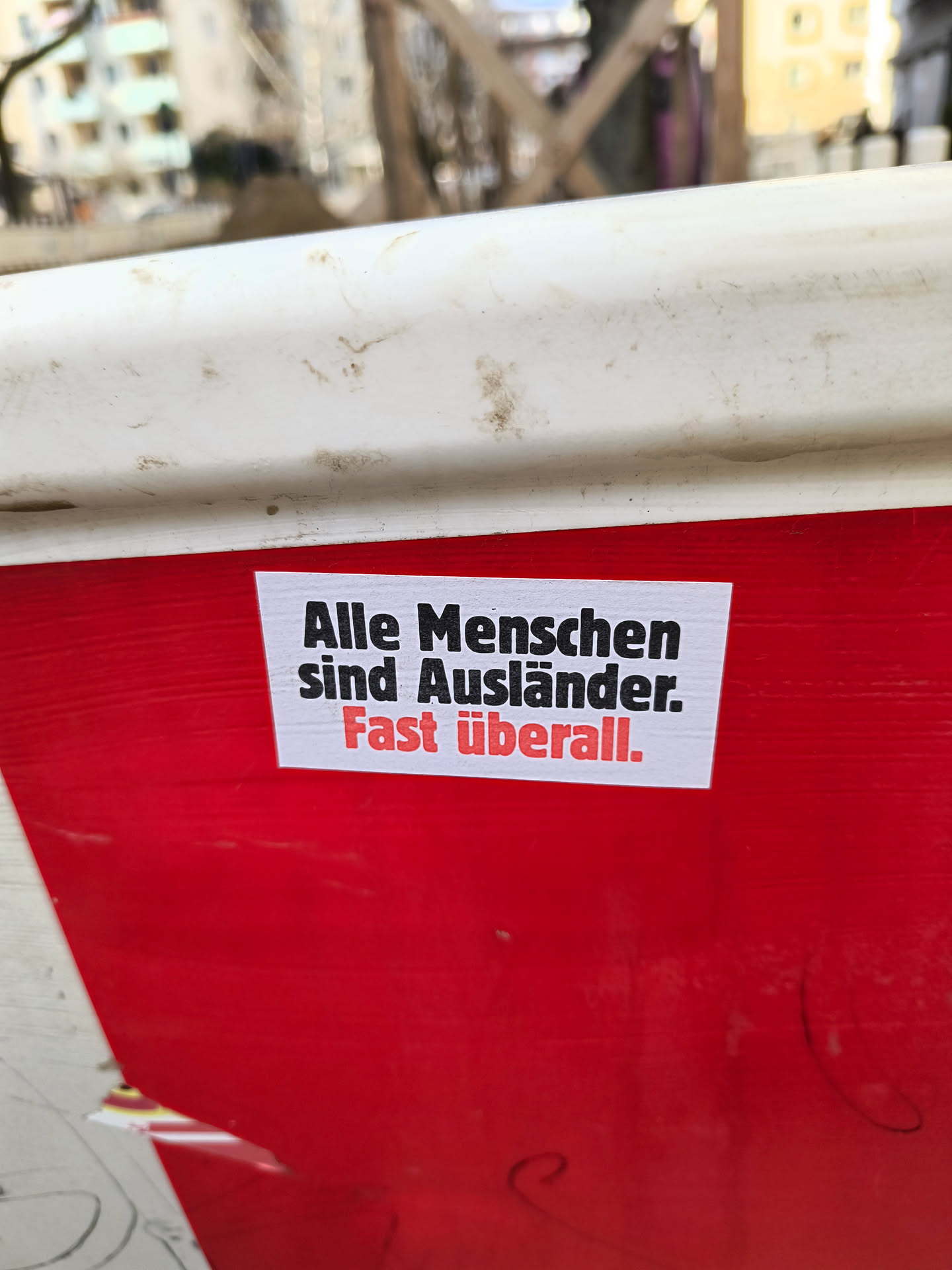 A sticker with the text "Alle Menschen sind Ausländer. Fast überall." meaning "All people are foreigners. Almost everywhere.", placed on a red and white background.