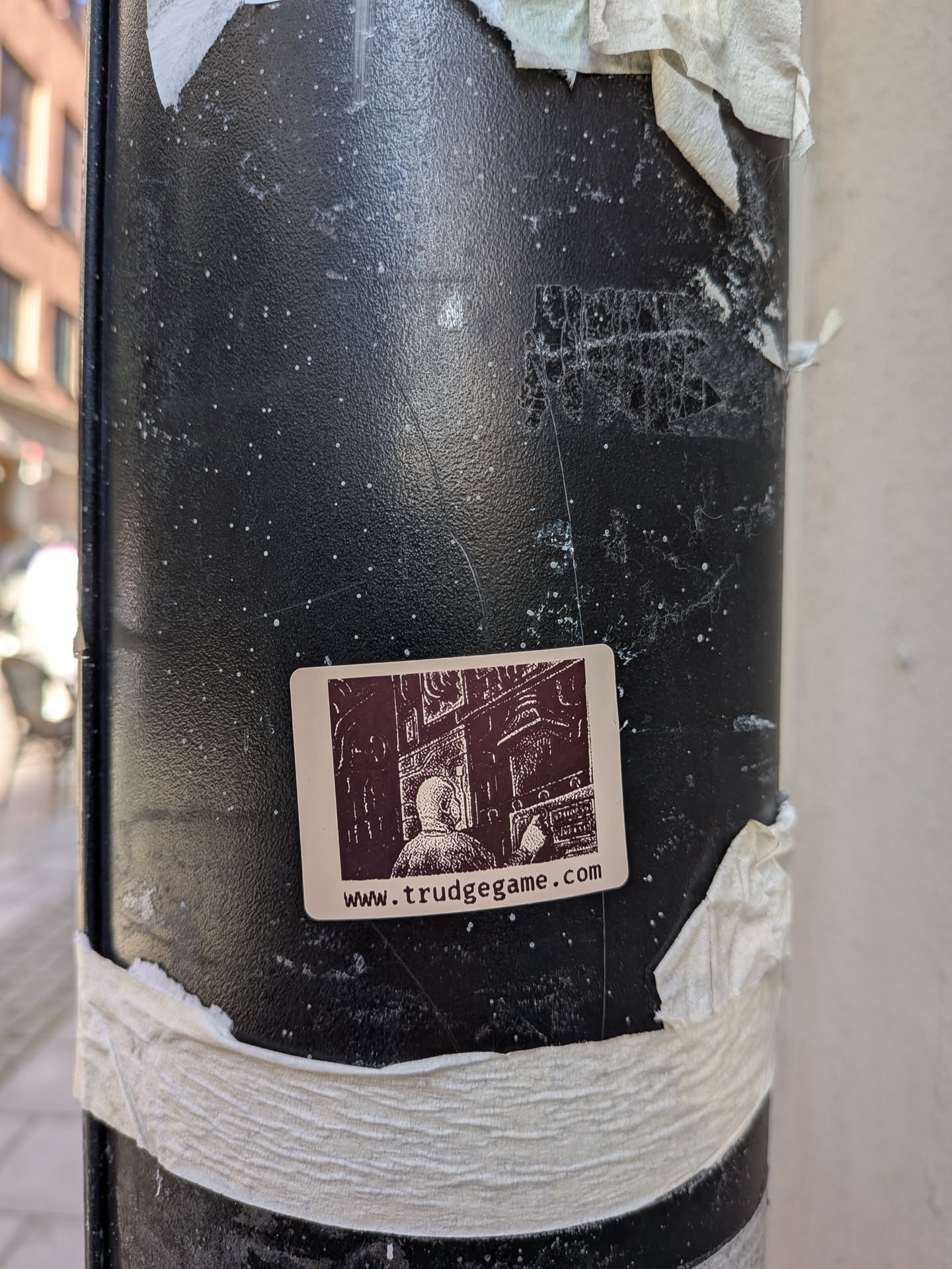 A rectangular sticker with rounded corners on a cream-colored background. It features a dark brown, detailed illustration in a woodcut or pixel-art style, depicting a hooded figure from behind looking at a complex control panel or machinery. Below the illustration, the website address 'www.trudgegame.com' is printed in a dark brown, lowercase font.