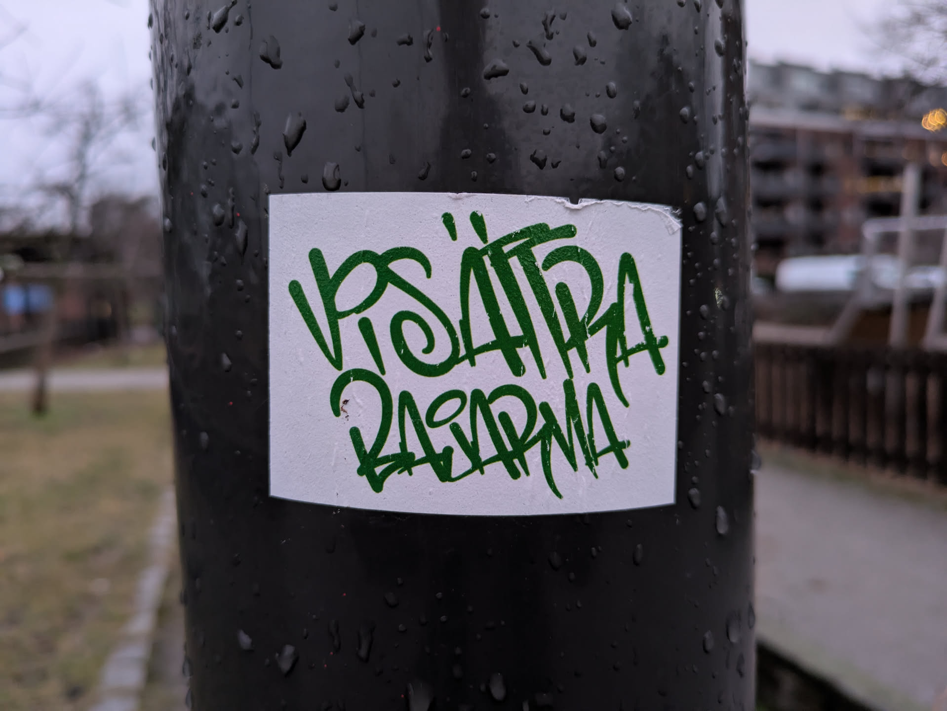 A close-up shot of a white sticker on a dark pole. The sticker features green graffiti-style lettering that appears to say "VISA HIRA RASARMA". The pole is wet, with water droplets visible on its surface. The background is slightly blurred but shows some outdoor scenery, such as trees and buildings in the distance.