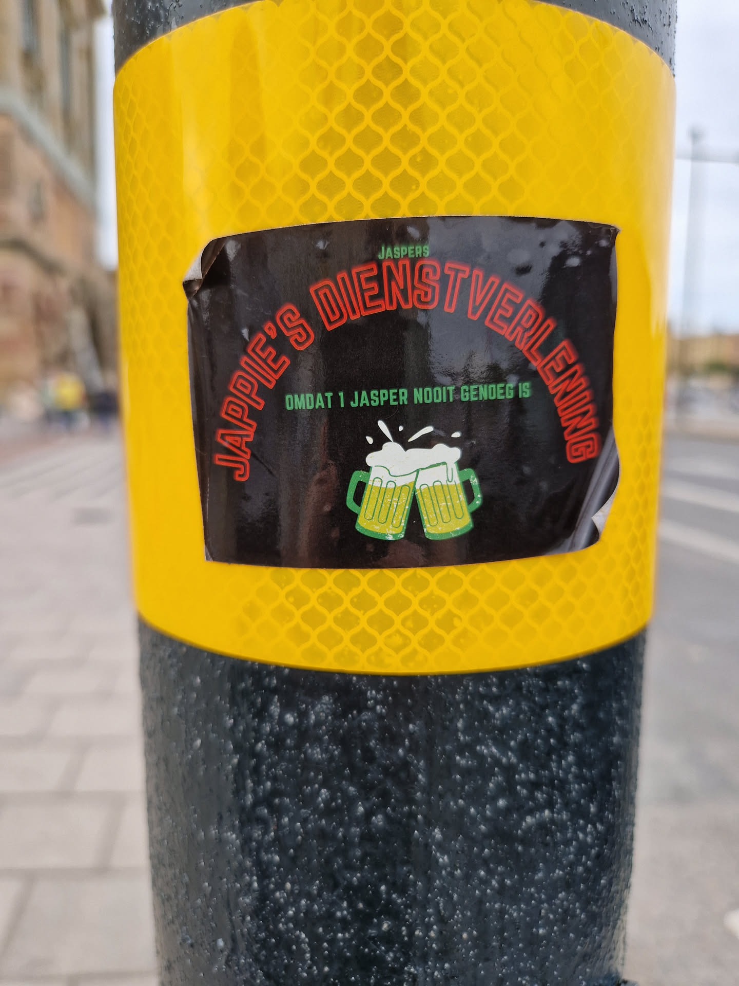A black sticker with green and red text on a yellow reflective surface. The text says "Jappie's Dienstverlening" and below it says "Omdat 1 Jasper nooit genoeg is." There is an image of two beer mugs. 