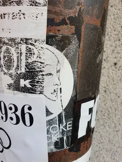 A black and white sticker with a circular design. The main focus is a stylized female face with a slightly abstract style.  The sticker is affixed to a rusty metal surface, which is covered in other stickers and markings.