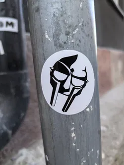 A round sticker featuring the iconic Metal Face mask of the legendary rapper MF DOOM. The sticker is black and white and is affixed to a metal pole.