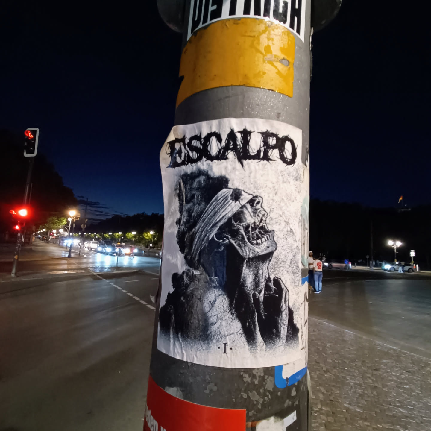 A black and white sticker featuring a skull-like figure with a blindfold, the word "ESCALPO" is visible at the top. The sticker is affixed to a pole, possibly a lamppost or signpost, on a city street at night. 