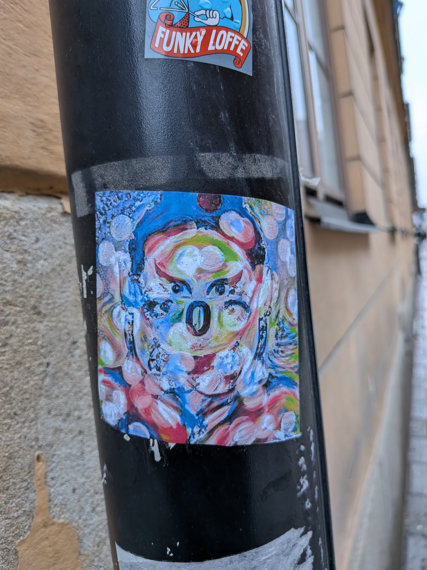 The sticker is square-shaped and features a highly stylized, abstract portrait of a face, possibly resembling a koala or bear due to its prominent, dark, oval-shaped nose. The artwork is rendered in a vibrant, painterly style with visible brushstrokes and textures, utilizing a rich palette of blues, pinks, reds, greens, yellows, and white. The eyes are small and dark, surrounded by intricate lines and multiple color layers, giving a somewhat distorted or multi-eyed appearance. The face is framed by numerous circular and oval shapes in various colors, creating a bubbly or textured aura against a background that blends blues and whites. The overall impression is one of a dynamic, colorful, and somewhat surreal artistic expression.