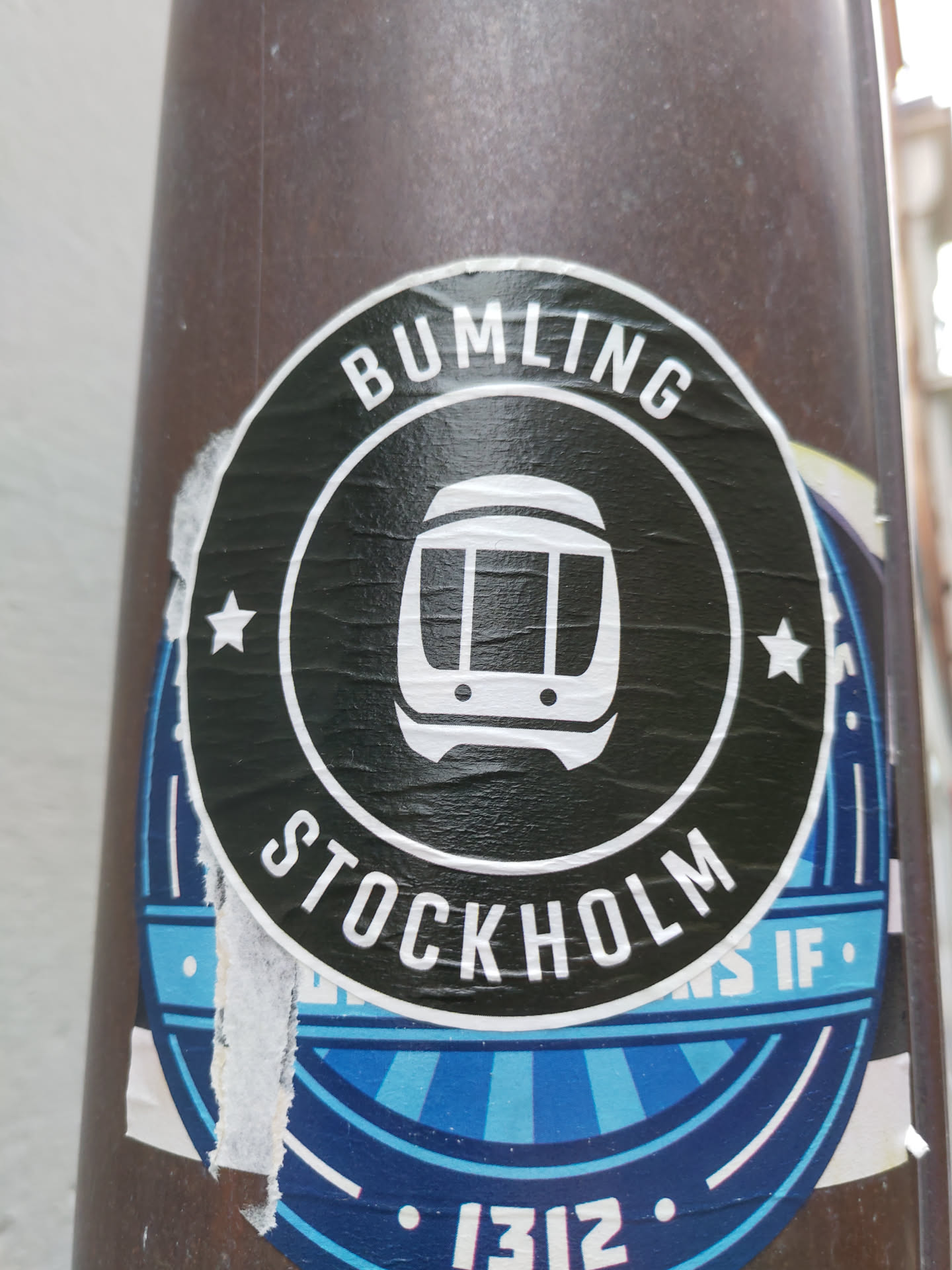 A black and white round sticker with the text "BUMLING" and "STOCKHOLM" in a circular pattern. There is an icon of a train in the center. The sticker is attached to a dark brown pole.