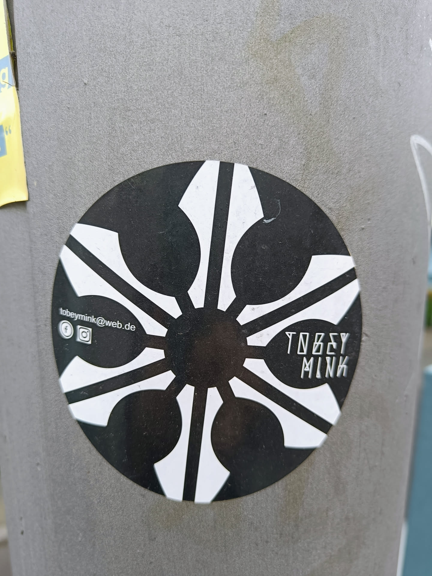 A round sticker featuring a striking black and white geometric design. The central motif resembles a stylized sun or flower, radiating outwards from a dark center.  The artist's name, TOBEY MINK, is subtly inscribed within the design. Social media links are included at the bottom.