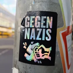 A sticker with a black background and iridescent text and graphic.  The text says "GEGEN NAZIS" (Against Nazis in German). The graphic depicts a fist crushing a swastika.