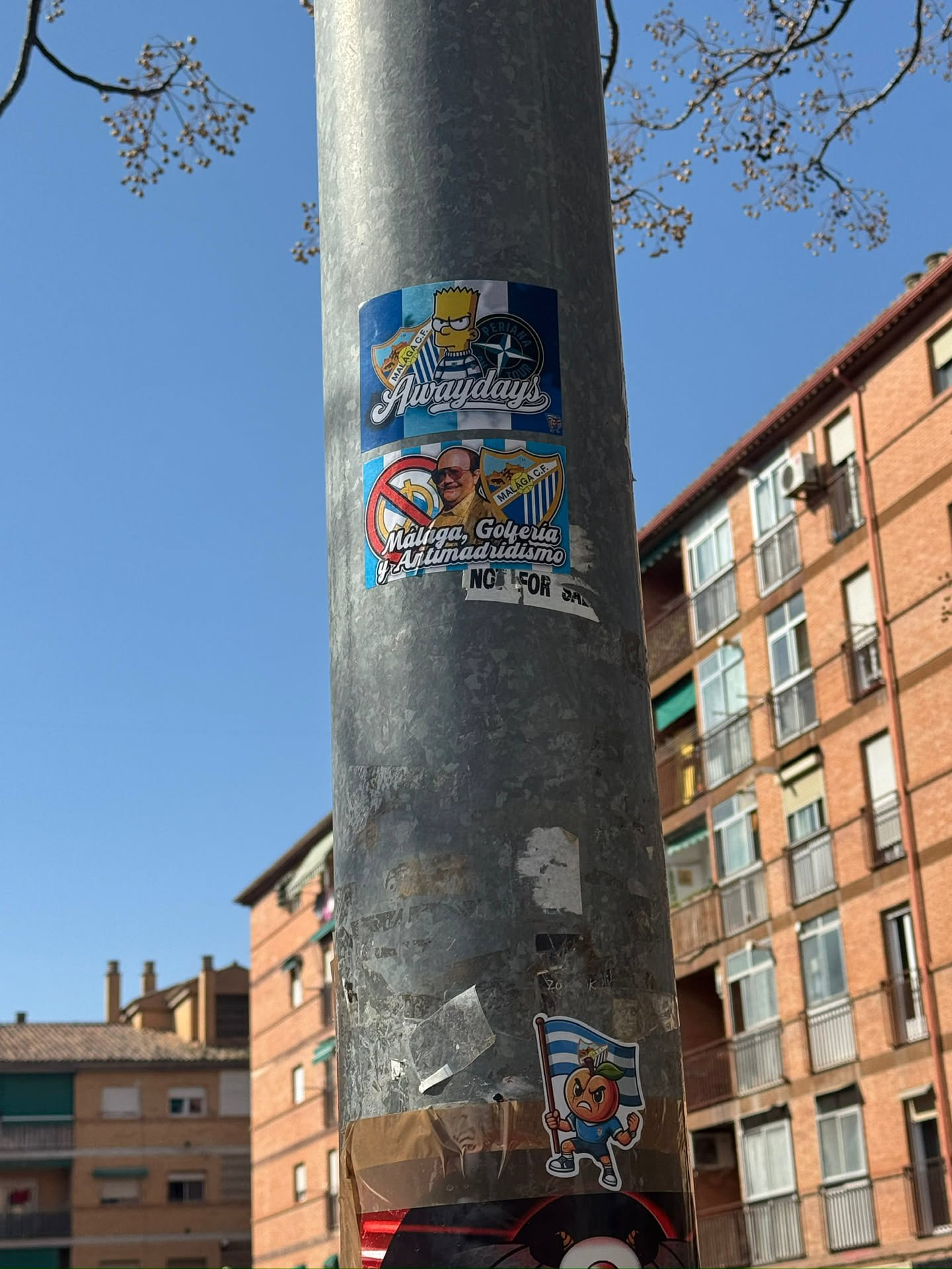The central sticker is a composite design, featuring two stacked rectangular sections. The upper section depicts Bart Simpson from The Simpsons, wearing a blue and white striped shirt, holding a flag with the 'MÁLAGA C.F.' crest and 'PERIANA TOUR' text. Below Bart, the word 'Awaydays' is prominently displayed in a stylized font, set against a light blue and white checkerboard background with a darker blue border. The lower section features a smiling man with glasses, flanked by a 'no money' symbol on the left and another 'MÁLAGA C.F.' crest on the right. Below the man, the text 'Málaga, Golfería y Antimadridismo' is written in white letters on a blue background. At the very bottom, in smaller white text, it reads 'NOT FOR SALE'. The sticker uses a palette of blues, white, yellow, red, and brown, and is adhered to a metal pole, showing some signs of wear.