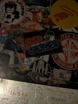 Street sticker A rectangular horizontal sticker set on a black background framed by a thin red outline. It represents the Lyon ultras group 'Bad Gones 1987'. The text 'BAD' is written in prominent distressed reddish-orange block letters, with 'DEPUIS 1987' printed in white underneath. To the right is the group's mascot—a snarling character wearing a black bowler hat—positioned directly above the stylized word 'GONES', which is textured with blue and dark blue patterned fill.