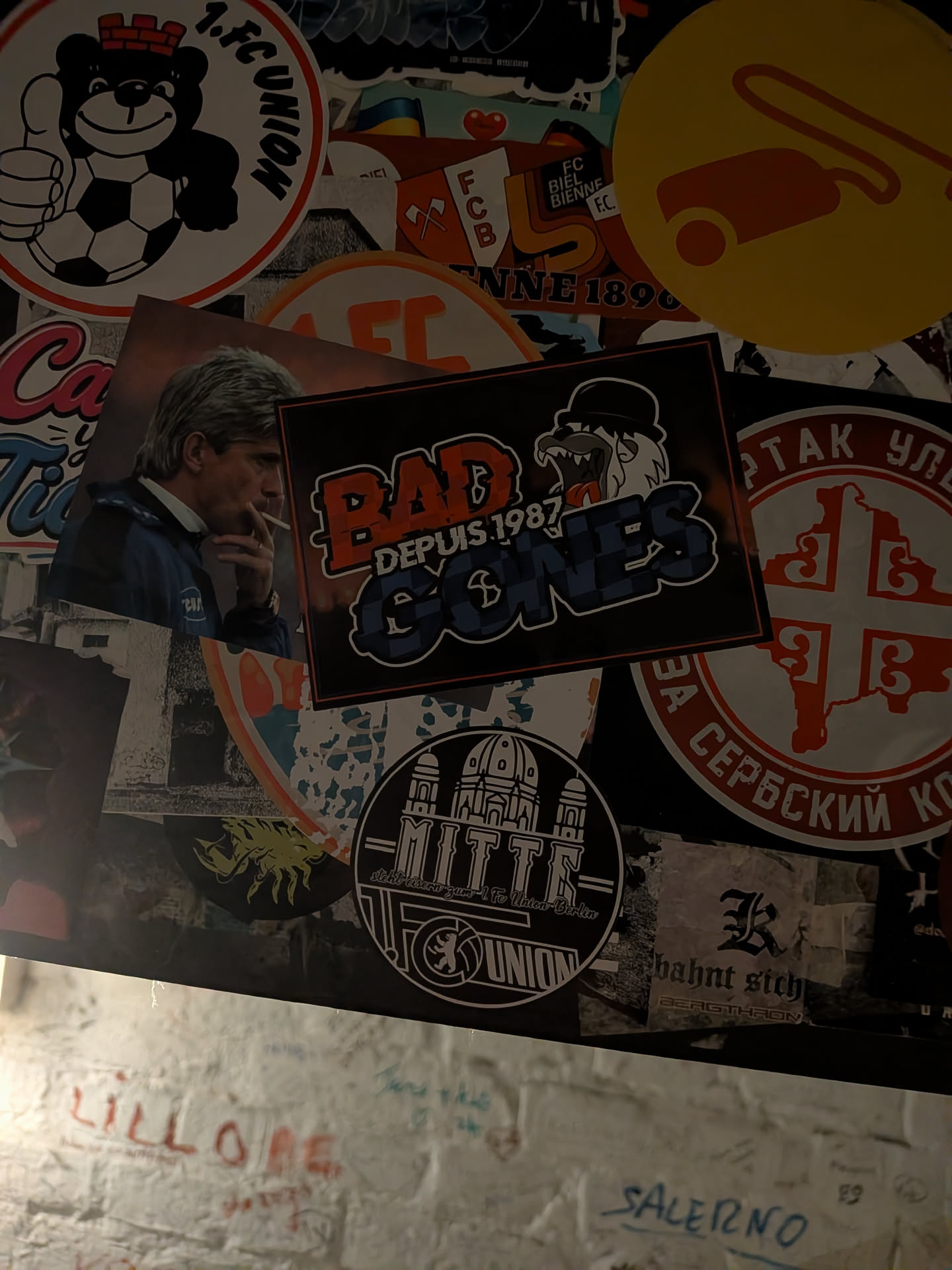 A rectangular horizontal sticker set on a black background framed by a thin red outline. It represents the Lyon ultras group 'Bad Gones 1987'. The text 'BAD' is written in prominent distressed reddish-orange block letters, with 'DEPUIS 1987' printed in white underneath. To the right is the group's mascot—a snarling character wearing a black bowler hat—positioned directly above the stylized word 'GONES', which is textured with blue and dark blue patterned fill.