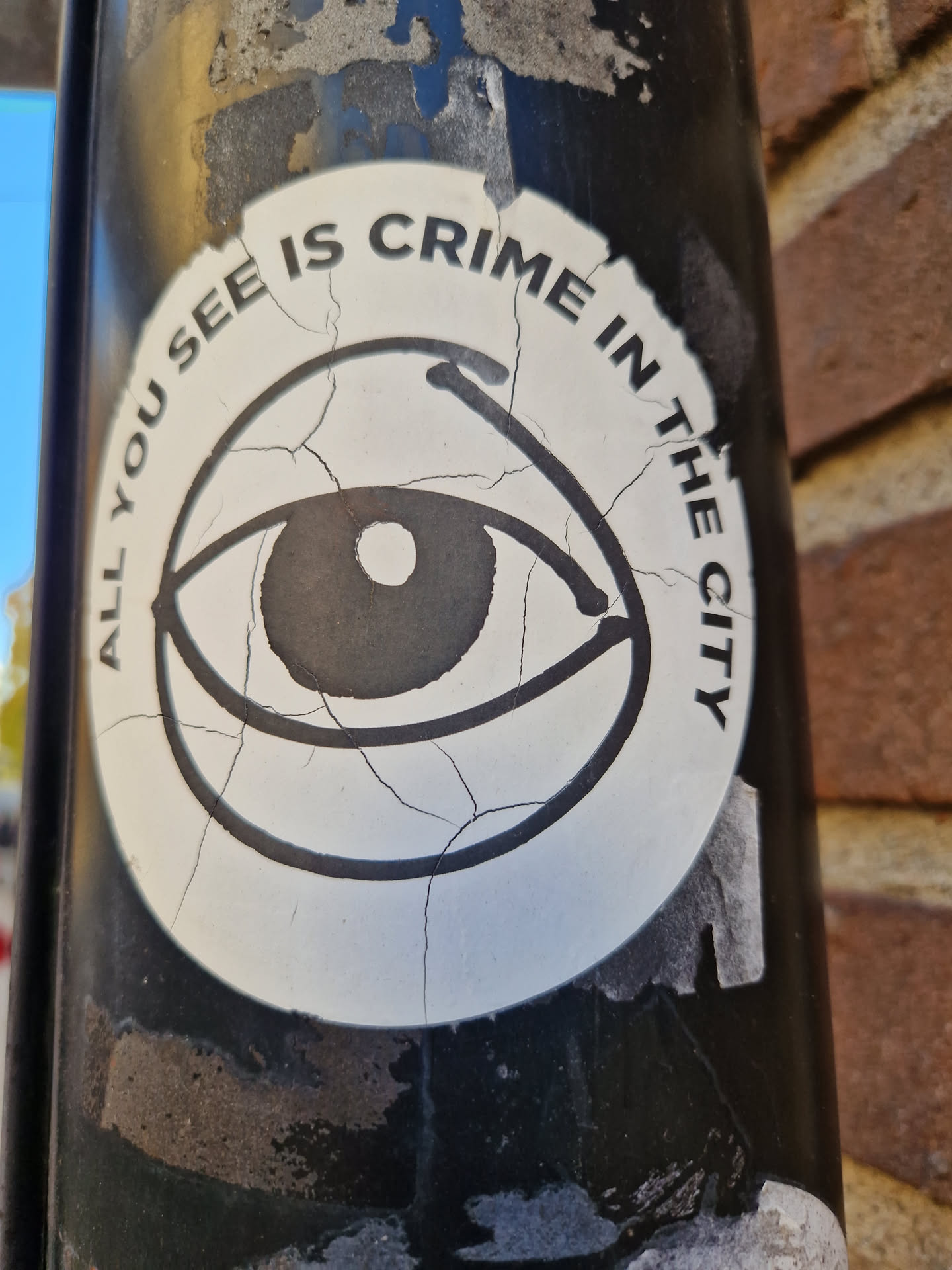 A worn sticker with an eye graphic and the text "All you see is crime in the city." The sticker shows signs of wear and tear, with cracks visible on its surface. The sticker is attached to a dark-colored pole.