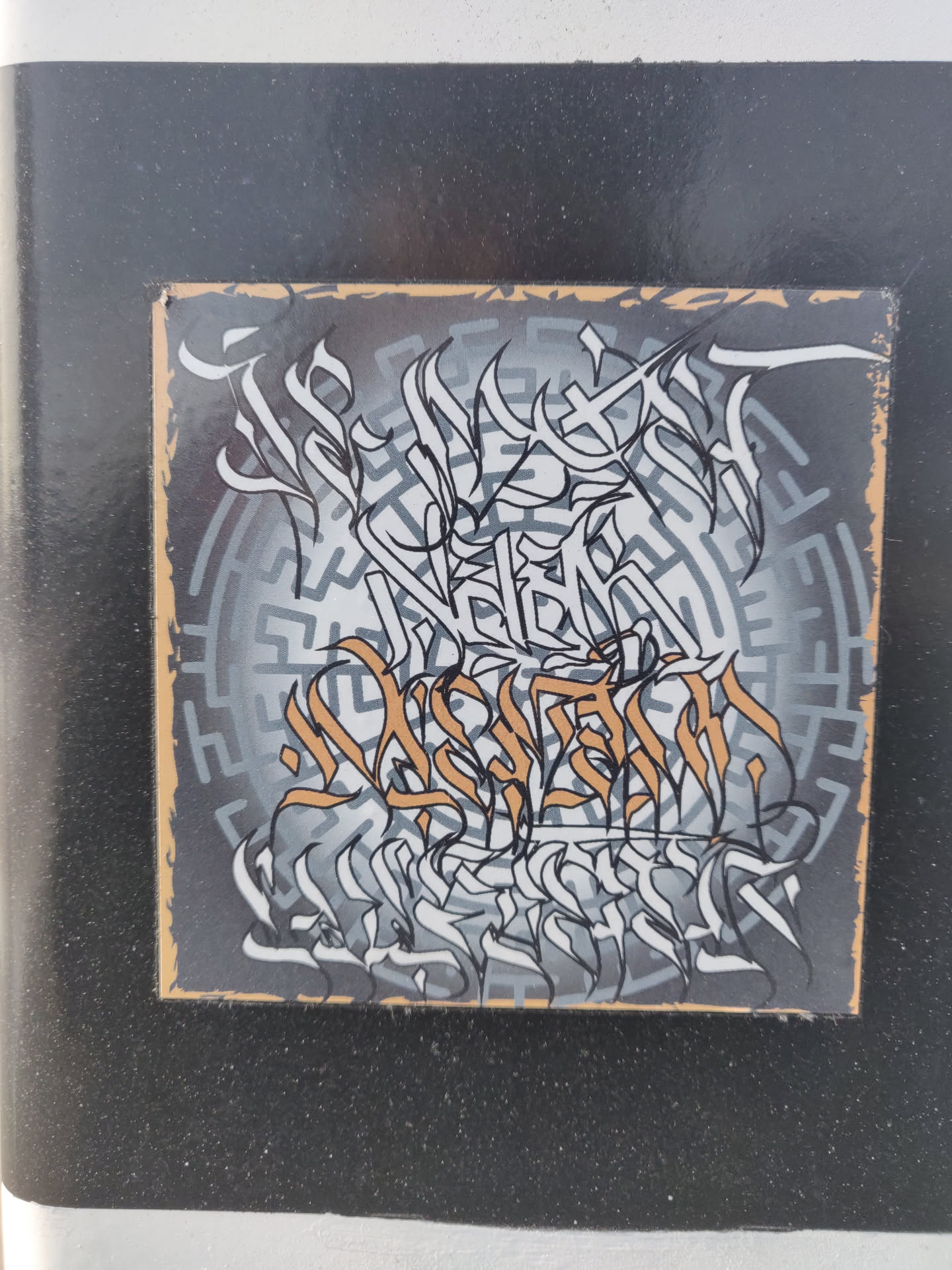 A square sticker featuring intricate black and white graffiti art on a gray background with a circular maze pattern.  Some elements of the art are highlighted in a muted gold color. The style is reminiscent of calligraphy or abstract lettering.