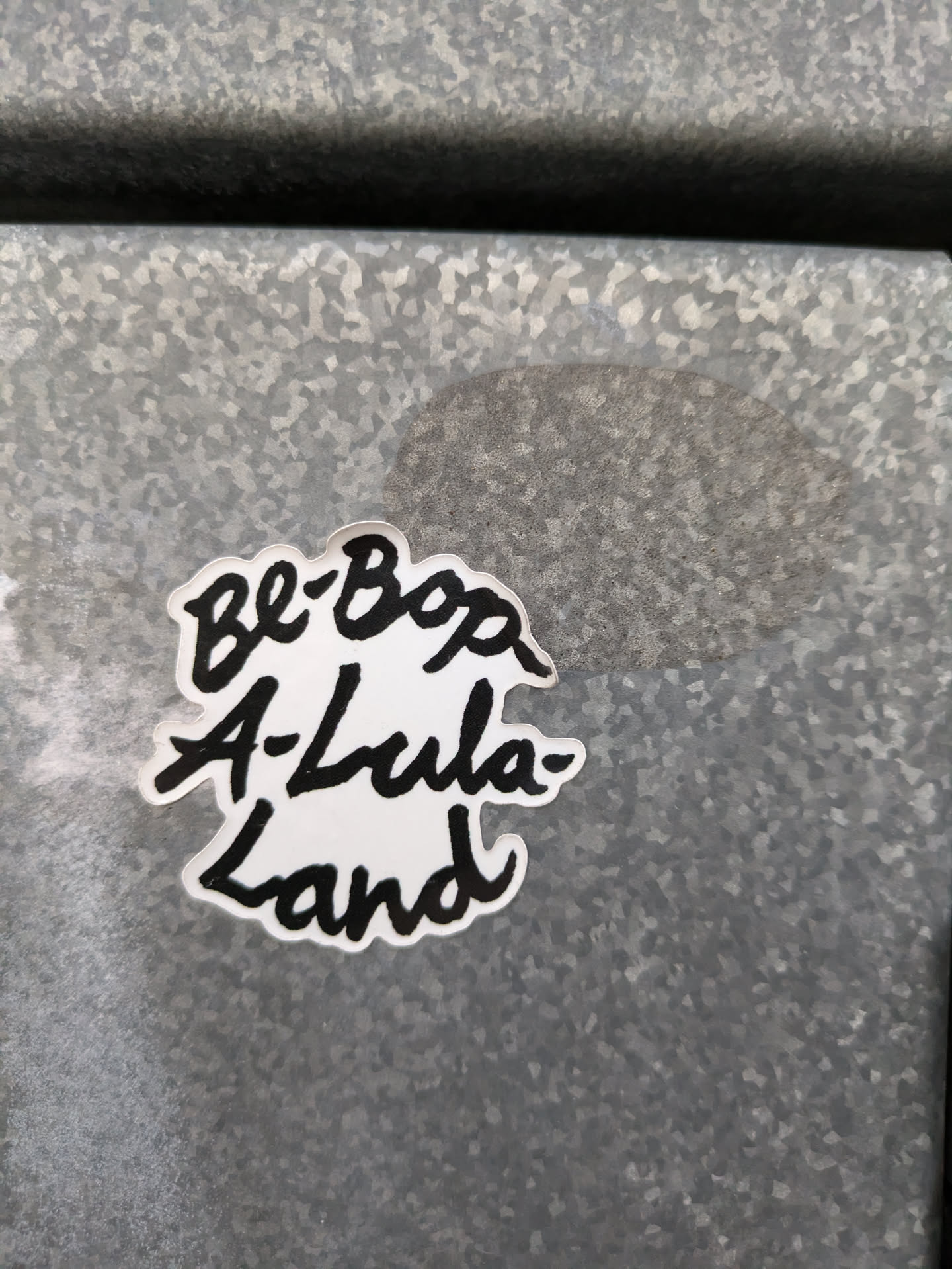 A white sticker with black text that says \"Be-Bop-A-Lula-Land\" in a handwritten style. The sticker is affixed to a gray metallic surface.