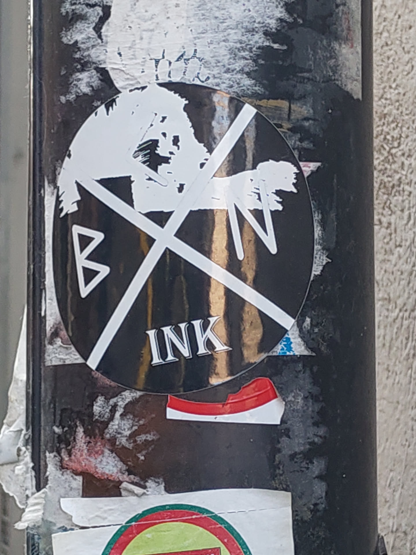 A round black and white sticker with an abstract design. It has two thick white lines crossing each other, with the letters "B" and "N" in the corners and a white brush-stroke-like shape within the circle. The word "INK" is printed in a serif font at the bottom. The sticker is stuck on a pole covered in other stickers.
