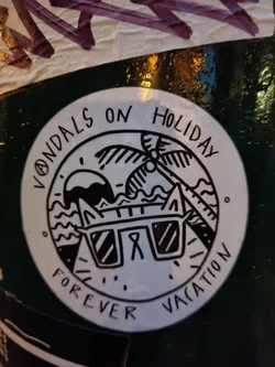 A round sticker with a black and white illustration of a cat wearing sunglasses in front of a beach scene. The text around the illustration says "Vandals on Holiday" at the top and "Forever Vacation" at the bottom.
