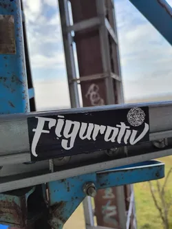 Black sticker with white lettering reading 'Figurativ'. It also contains a small QR code. The sticker is affixed to a rusty blue metal structure.
