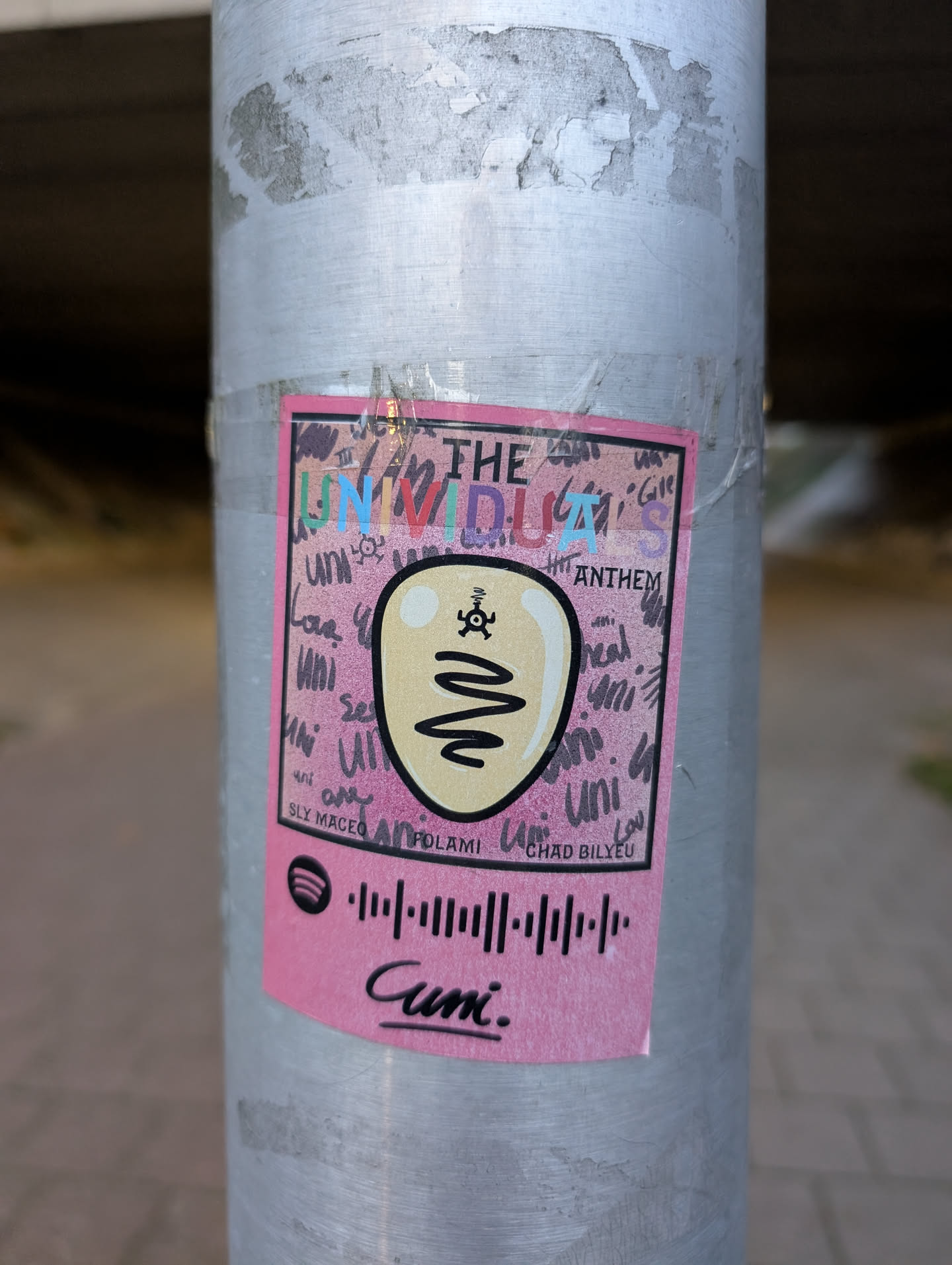 A rectangular sticker with a bright pink background, outlined in black, affixed to a metal pole. The sticker features a central, light yellow/beige oval shape with a thick black outline, resembling a stylized head or face. Above this shape is a small black, multi-limbed symbol, and below it, a thick, wavy black line acts as a mouth. A small white highlight is visible on the upper left of the yellow shape. The top of the sticker reads 'THE' in black, followed by 'UNIVIDUAL' in multi-colored (blue, green, purple, red, orange) block letters, and 'ANTHEM' in black. The background is filled with repeated, scrawled instances of the word 'uni' and other faint scribbles in black. Below the central graphic, the names 'SLY MACEO', 'ANPOLAMI', and 'CHAD BILYEU' are listed in black. The bottom section prominently displays a black Spotify code (a soundwave pattern) and below it, the word 'Cuni.' is written in a stylized black script font. The sticker shows signs of wear and is partially covered with clear tape.