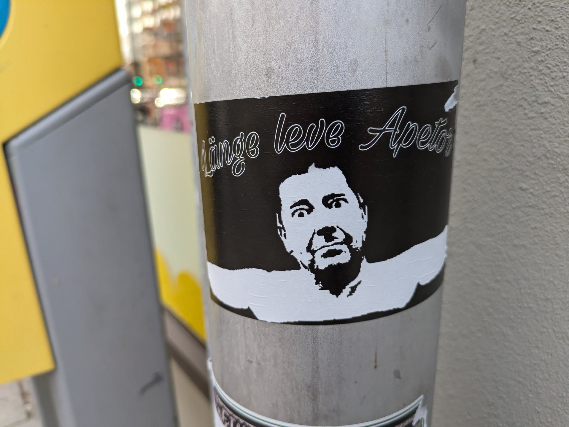 This sticker shows a stylized black and white portrait of a man's face with a surprised or slightly fearful expression, set against a black background.  Above the portrait is the phrase "Linge leve Apetos" written in a cursive script.