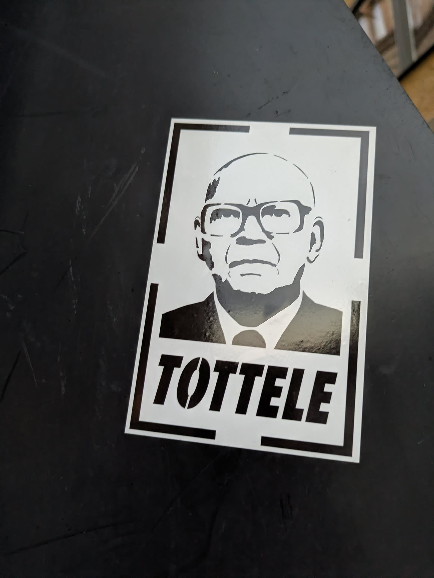 The sticker features a black and white stencil portrait of a bald man wearing glasses and a suit. The portrait is rendered with simple, bold lines, and the text 'TOTTELLE' is prominently displayed in stenciled capital letters below the image. The sticker has a white background with a thin black border, and a thicker black border framing the entire design.
