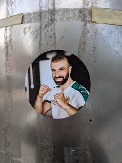 The sticker is circular, depicting a smiling man with a dark beard and short, styled hair. He is shown from the chest up, wearing a white sports jersey with green accents on the shoulders and sleeves. His hands are raised in a boxing or fighting stance, with fists lightly clenched. A gold and black crest-like logo is visible on the left side of his chest, and text like 'PUMA' and 'INTERSPORT' can be partially seen on his shirt. The background behind him is dark and slightly blurred, suggesting an indoor setting like a gym.