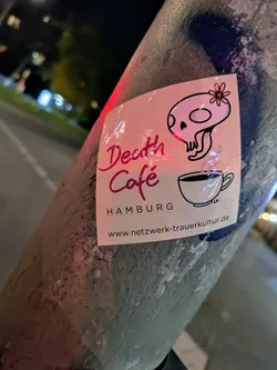 A sticker for Death Cafe in Hamburg. It features a cute skull illustration and a cup of coffee. The website is printed on the sticker.