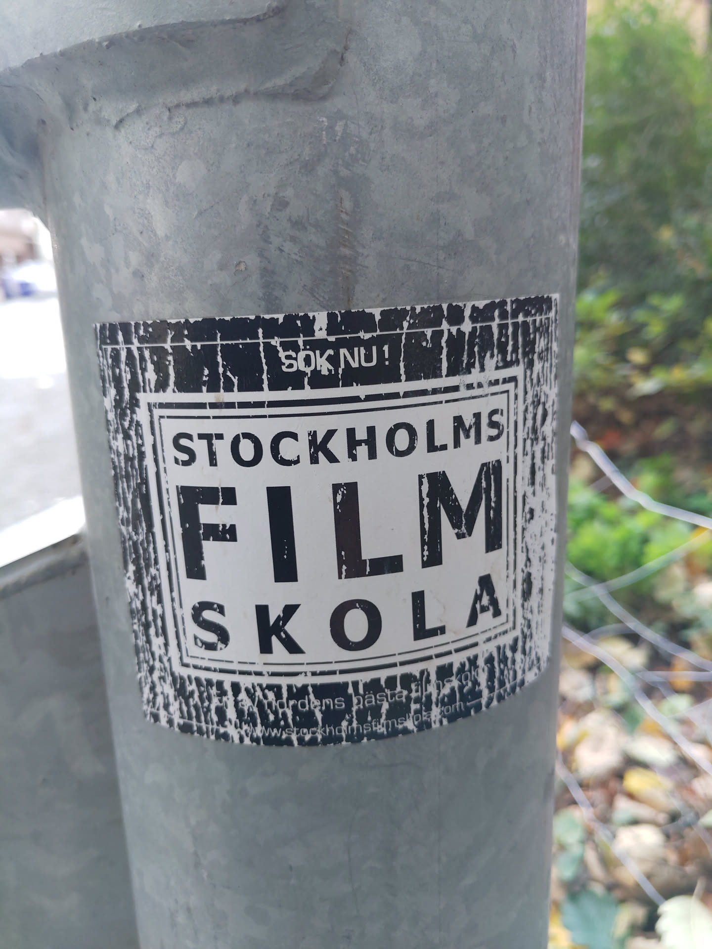 A worn sticker advertising Stockholm Film Skola.  The text 'Stockholms Film Skola' is prominently displayed in a distressed, vintage-style font. A website address is partially visible at the bottom.
