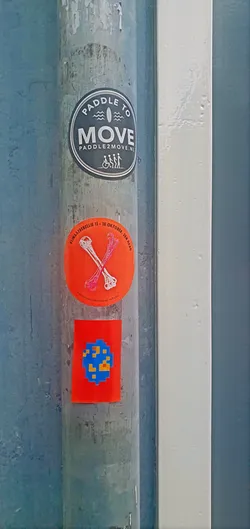 A photo of three stickers on a metal pole. The top sticker is round and black with the words "PADDLE TO MOVE PADDLE2MOVE.NL" and a graphic of a surfer and some figures in wheelchairs. The middle sticker is round and orange with the words "KLIMAATREBELLIE 11 - 18 OKTOBER, DEN HAAG" and an image of crossed bones. The bottom sticker is square and red with blue and yellow pixel art.