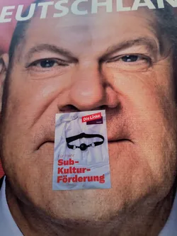 A sticker placed over the mouth of a politician in a poster.  The sticker depicts a ball gag and the text 'Für mehr Subkulturförderung' which translates to 'For more subculture funding'.