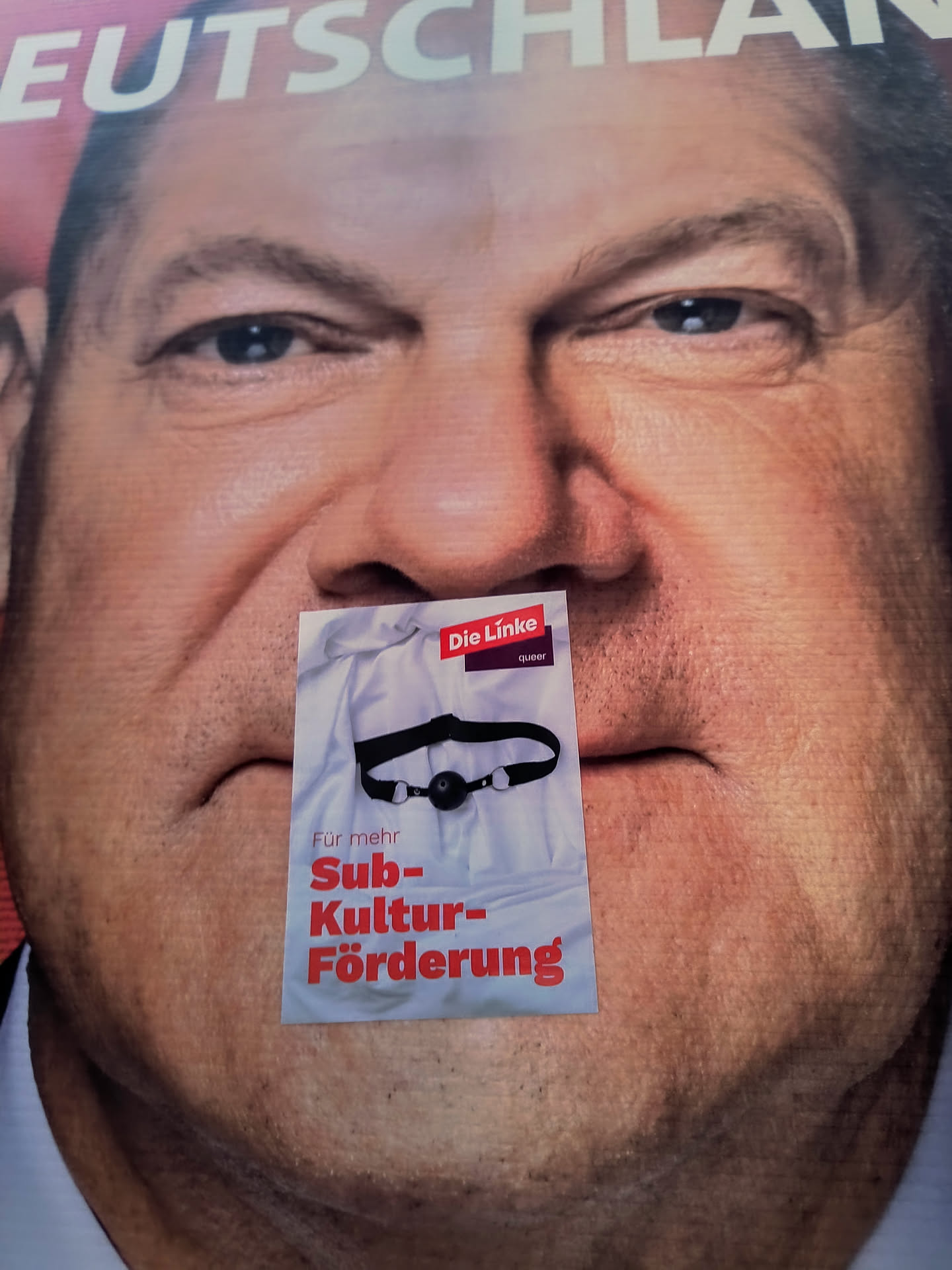 A sticker placed over the mouth of a politician in a poster.  The sticker depicts a ball gag and the text 'Für mehr Subkulturförderung' which translates to 'For more subculture funding'.