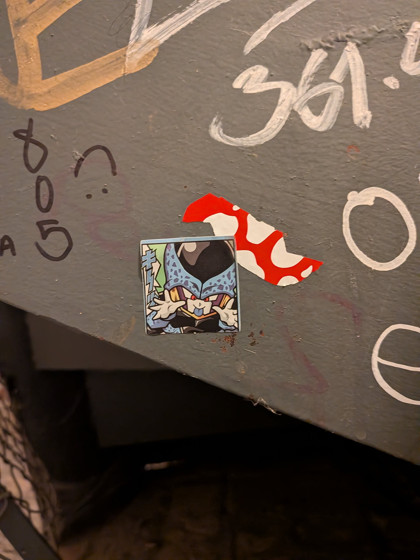A square sticker depicting the character Cell Jr. from Dragon Ball Z in a chibi/stylized art style. The character is light blue with black spots, red eyes, and dark purple/black accents, sticking its tongue out and waving its hands playfully next to its face. Japanese manga sound effect text appears vertically along the left edge against a pale green background.