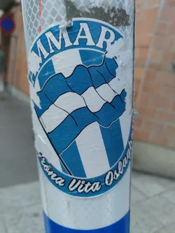 A weathered sticker featuring a blue and white crest with the words "Fröna Vita Osla" and what looks like the team's name partially visible above. The sticker is affixed to a pole and shows signs of wear, adding to its character.