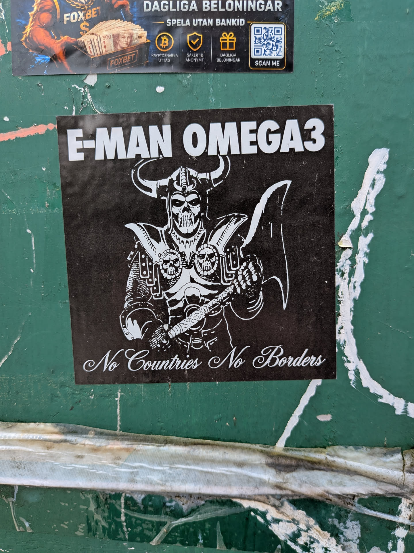 A square black sticker with white graphics and text, pasted on a green surface. At the top, the words 'E-MAN OMEGA3' are printed in a bold, white, sans-serif typeface. The central illustration depicts a skeletal warrior in detailed armor, wearing a horned helmet and holding a large battle axe. The armor features skull designs on the shoulder plates. At the bottom of the sticker, the phrase 'No Countries No Borders' is written in an elegant, white cursive script.