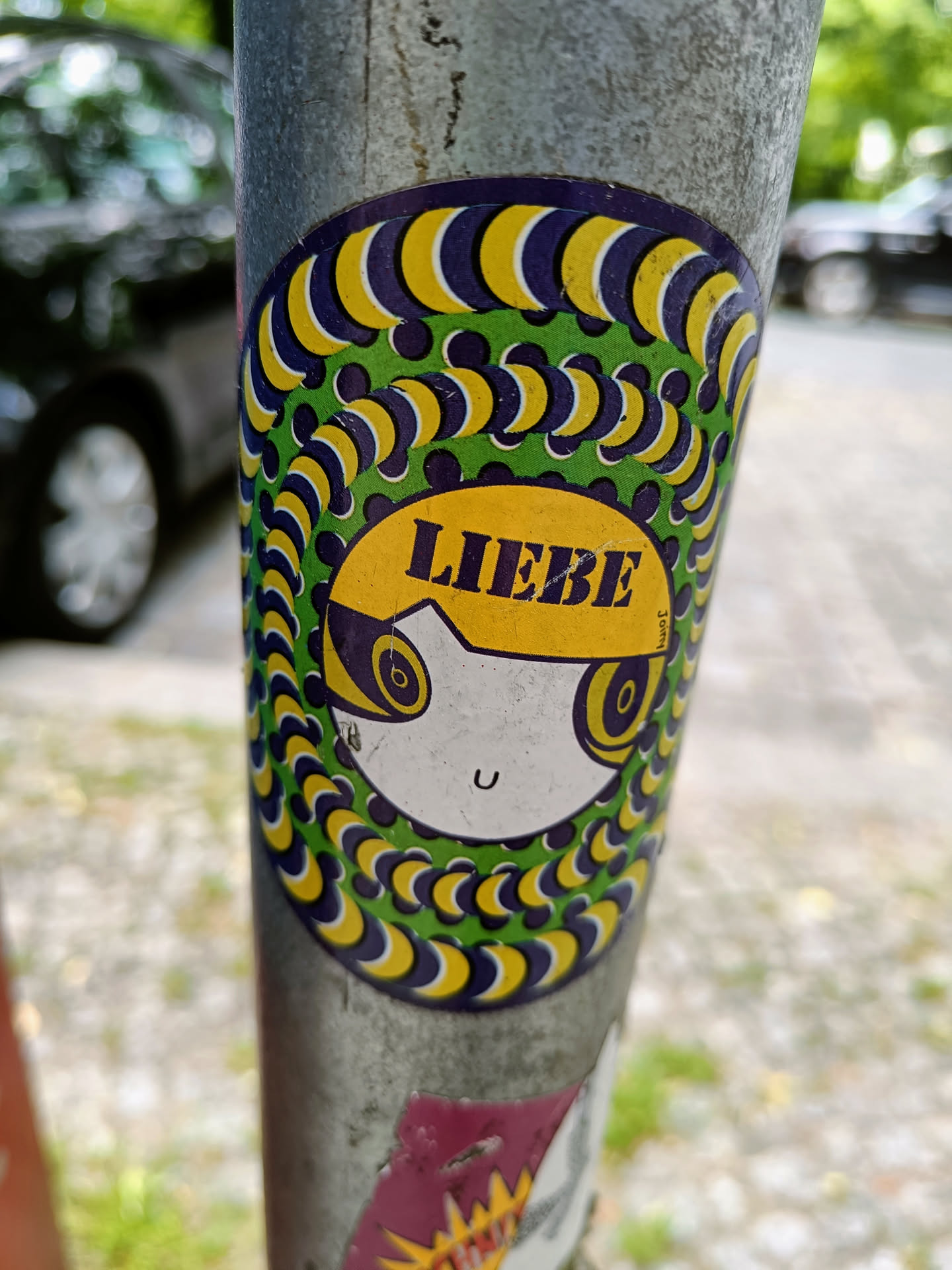 A round sticker featuring a cartoon face with the word "Liebe" (German for Love) on its forehead. The face is surrounded by a mesmerizing pattern of yellow and purple crescent shapes on a green background, creating an optical illusion effect. The sticker is affixed to a metal pole.