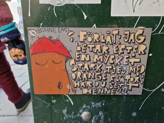 The sticker shows a simple drawing of an orange character with a red hat. The text around it says "Sorry, I'm looking for a very strong girl with orange floaters! Have you seen her?"