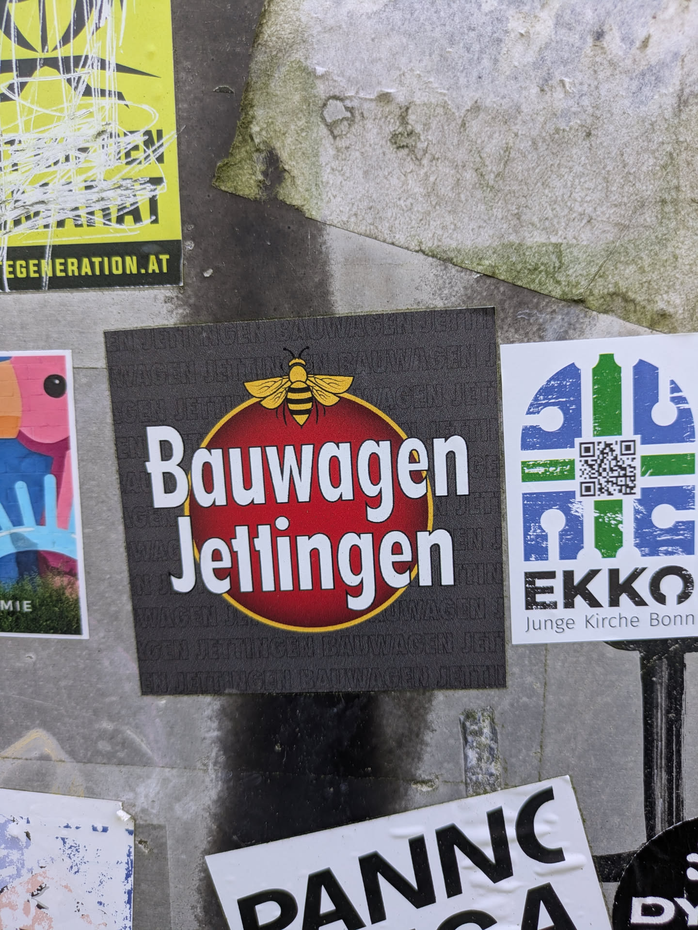 A square, dark grey sticker centered on a wall, featuring a prominent red circle with a thin yellow outline. Inside the circle, the words "Bauwagen Jettingen" are written in bold white letters. A yellow bee graphic is positioned directly above the red circle. The dark grey background of the sticker is subtly patterned with faint, repeating text that also reads "BAUWAGEN JETTINGEN".