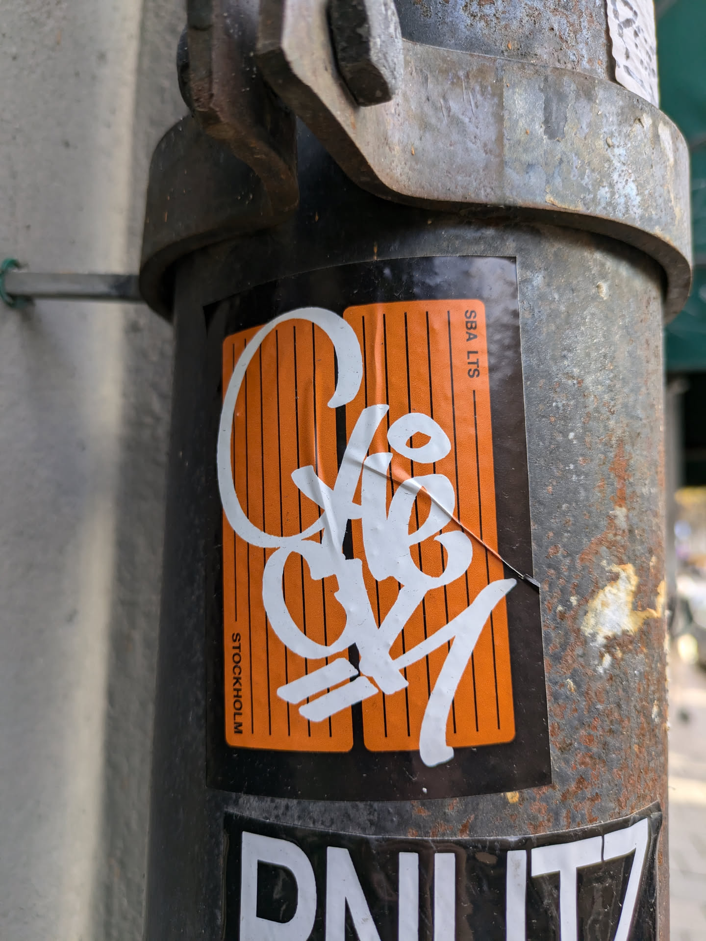 A close-up shot of an orange and white sticker affixed to a rusty metal pole. The sticker features stylized white graffiti-style lettering on an orange background.  The word "Stockholm" is visible at the bottom of the sticker, suggesting a possible origin or artist location.